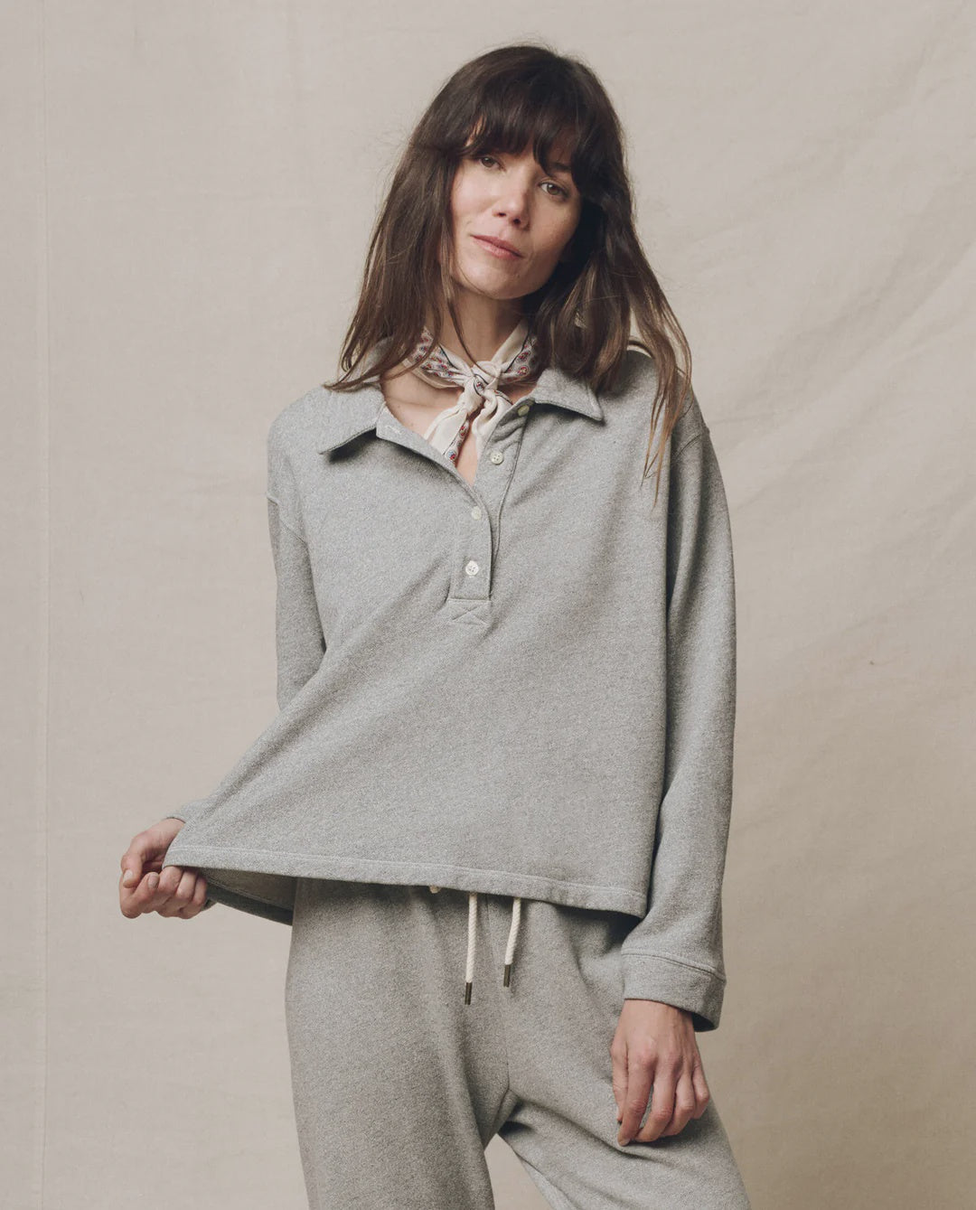 A woman with long brown hair wears The Great Inc.'s Rugby Sweater and matching pants, posing against a neutral background while holding out the bottom of her sweater. She accessorizes with a light-colored scarf tied around her neck.