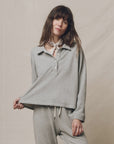 A woman with long brown hair wears The Great Inc.'s Rugby Sweater and matching pants, posing against a neutral background while holding out the bottom of her sweater. She accessorizes with a light-colored scarf tied around her neck.