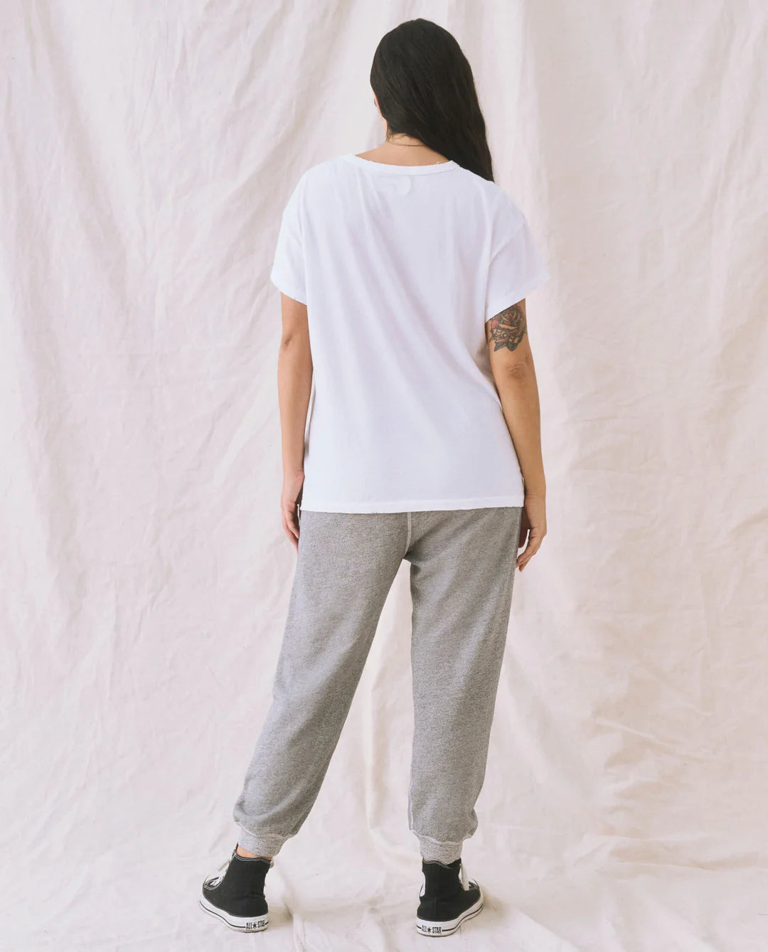 A person faces away, wearing a loose white T-shirt, The Great Inc. Cropped Sweat Pant in washed cotton terry, and black sneakers. Their long dark hair falls to one side, exposing a tattoo on their upper left arm against a light textured fabric backdrop.