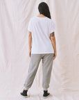A person faces away, wearing a loose white T-shirt, The Great Inc. Cropped Sweat Pant in washed cotton terry, and black sneakers. Their long dark hair falls to one side, exposing a tattoo on their upper left arm against a light textured fabric backdrop.