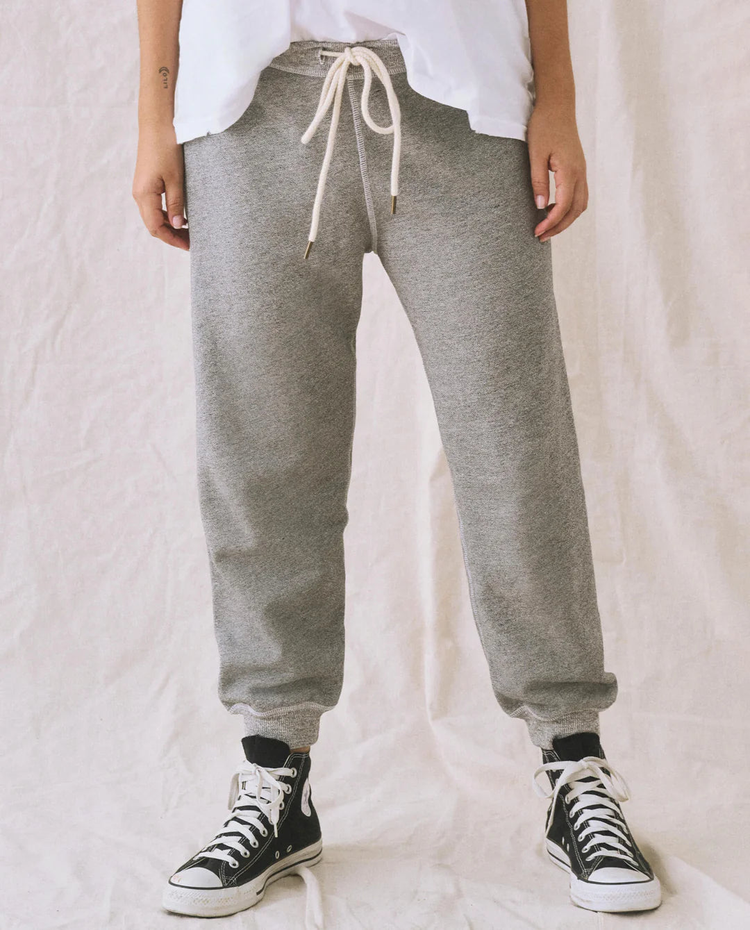 A person models The Great Inc. Cropped Sweat Pant in grey washed cotton terry with a white drawstring, paired with a white shirt and black high-top sneakers, standing against a light-colored fabric backdrop. Their head is not visible in the photo.