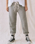 A person models The Great Inc. Cropped Sweat Pant in grey washed cotton terry with a white drawstring, paired with a white shirt and black high-top sneakers, standing against a light-colored fabric backdrop. Their head is not visible in the photo.