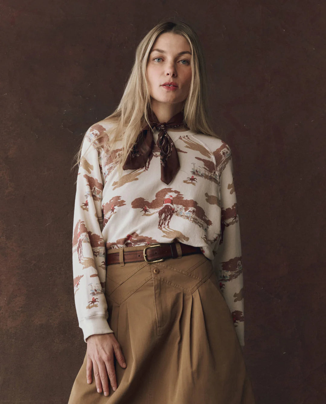 A woman with long blonde hair wears The Slouch Sweatshirt by The Great Inc., styled with a brown scarf and a tan high-waisted skirt. She stands against a dark brown backdrop, gazing to the side for a timeless, vintage appeal.