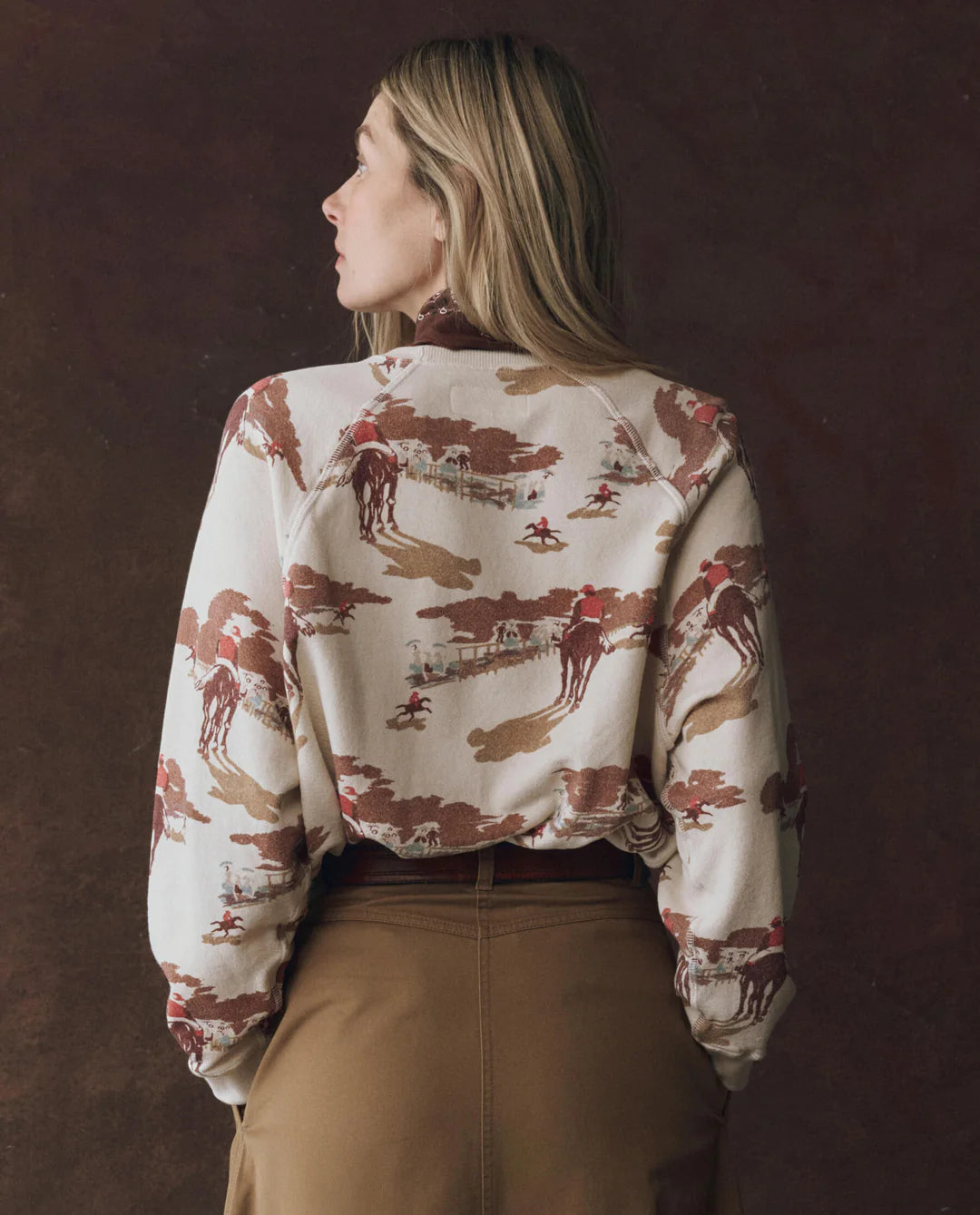 A woman with long blonde hair faces away, wearing The Great Inc.'s The Slouch Sweatshirt in vintage cream and brown with Western illustrations, paired with high-waisted brown pants, set against a dark brown background.
