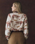 A woman with long blonde hair faces away, wearing The Great Inc.'s The Slouch Sweatshirt in vintage cream and brown with Western illustrations, paired with high-waisted brown pants, set against a dark brown background.