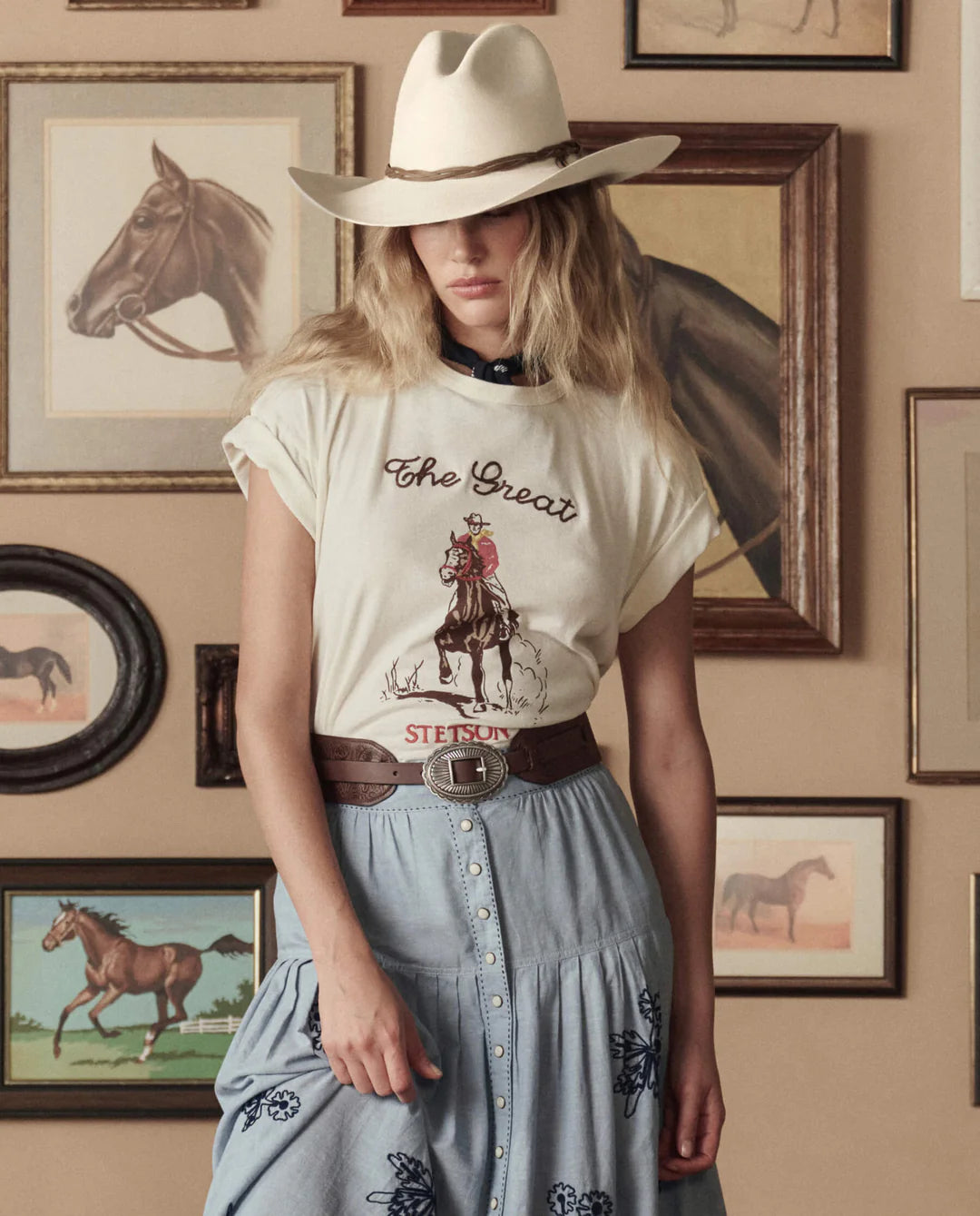 A woman in a cowboy hat, graphic tee, denim skirt, and The Great Inc. Tooled Waist Belt stands before a wall of horse art, her head tilted so the hat shades part of her face.