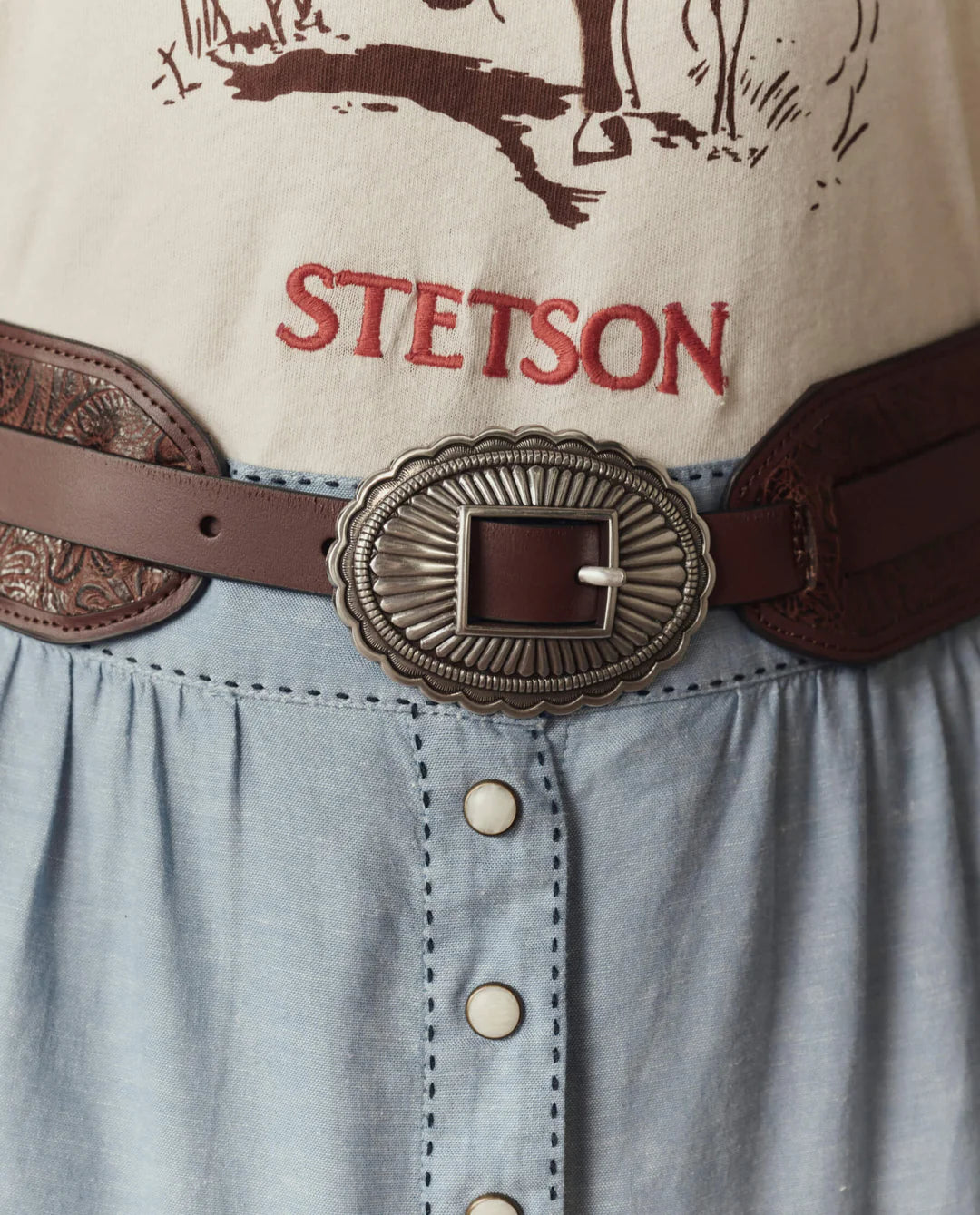 A close-up of a person in a beige shirt with "STETSON" in red, wearing The Great Inc.'s Tooled Waist Belt with an ornate silver buckle, paired with a light blue buttoned skirt.