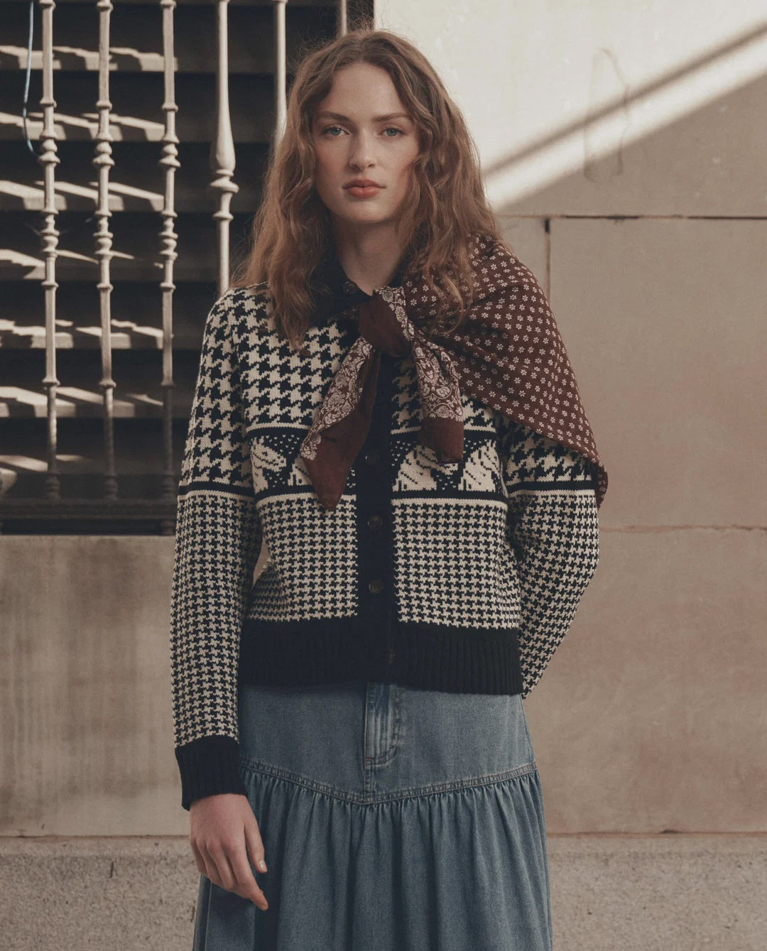 The Pony Cardigan in soft knit, styled with a denim skirt and scarf, beside a rustic stone wall and railing.