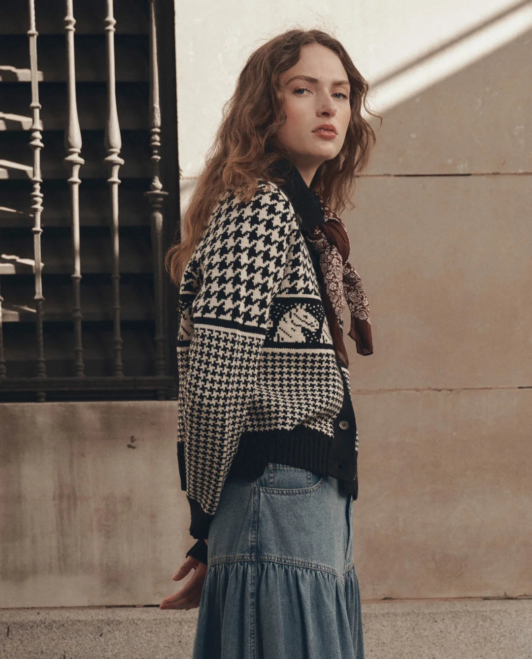 The Pony Cardigan by The Great Inc. styled with a long denim skirt and scarf beside a beige wall and window.