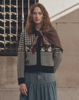 The Pony Cardigan in soft knit, styled with a denim skirt and scarf, beside a rustic stone wall and railing.
