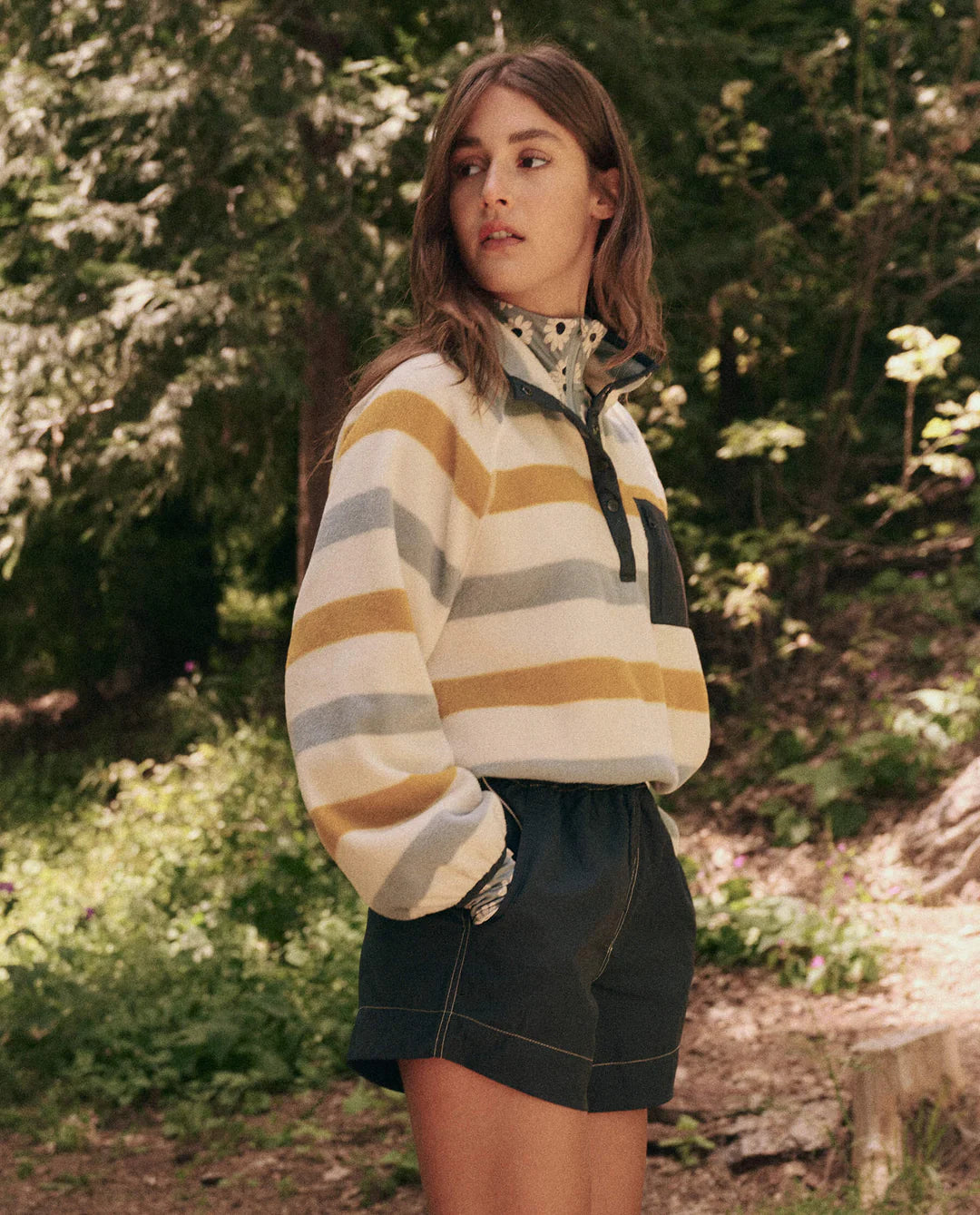 In a forest, a long-haired person embodies adventure in The Great Inc.'s Excursion Pullover striped in white, grey, and mustard. Paired with dark shorts, their technical attire blends perfectly into the trees and greenery as they gaze sideways.