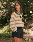 In a forest, a long-haired person embodies adventure in The Great Inc.'s Excursion Pullover striped in white, grey, and mustard. Paired with dark shorts, their technical attire blends perfectly into the trees and greenery as they gaze sideways.