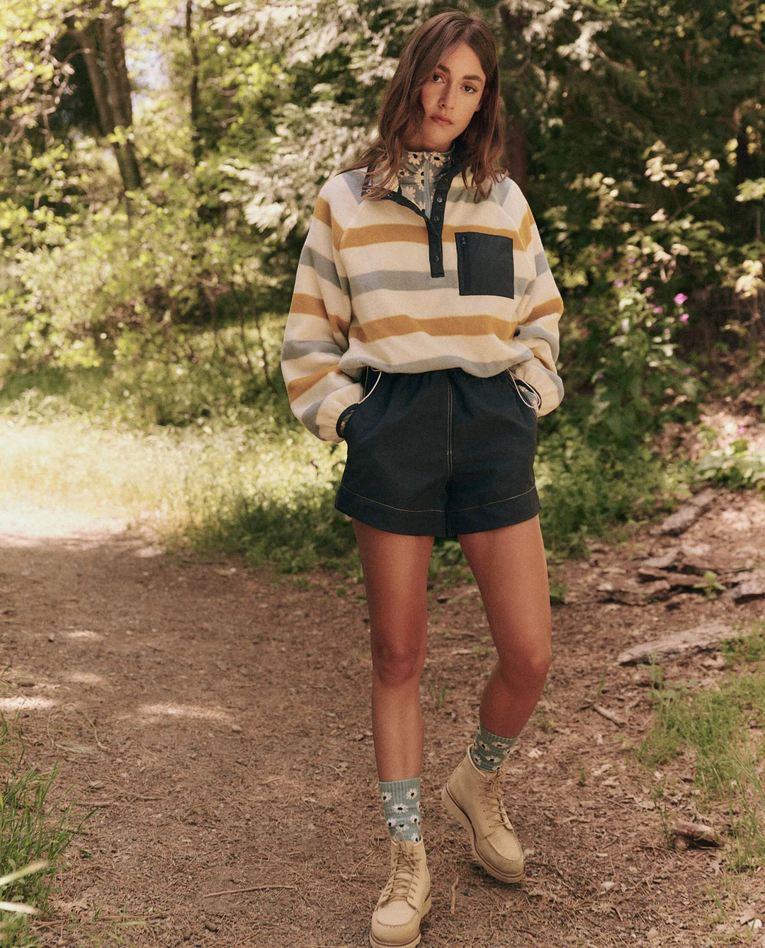 A woman on a wooded path embraces nature, wearing a striped pullover with a dark pocket, high-waisted &quot;The Rover Short&quot; by The Great Inc., patterned socks, and light boots. Her hands rest in her pockets as she&#39;s surrounded by trees and greenery.