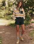 A woman on a wooded path embraces nature, wearing a striped pullover with a dark pocket, high-waisted "The Rover Short" by The Great Inc., patterned socks, and light boots. Her hands rest in her pockets as she's surrounded by trees and greenery.