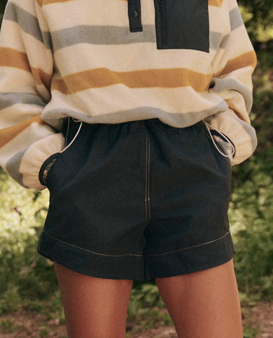 Someone is wearing The Rover Short by The Great Inc., which are dark, high-waisted, water-repellant shorts with pockets. They're paired with a striped long-sleeve top in yellow, gray, and beige hues amidst a backdrop of greenery, highlighting practical outdoor wear.