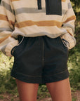 Someone is wearing The Rover Short by The Great Inc., which are dark, high-waisted, water-repellant shorts with pockets. They're paired with a striped long-sleeve top in yellow, gray, and beige hues amidst a backdrop of greenery, highlighting practical outdoor wear.