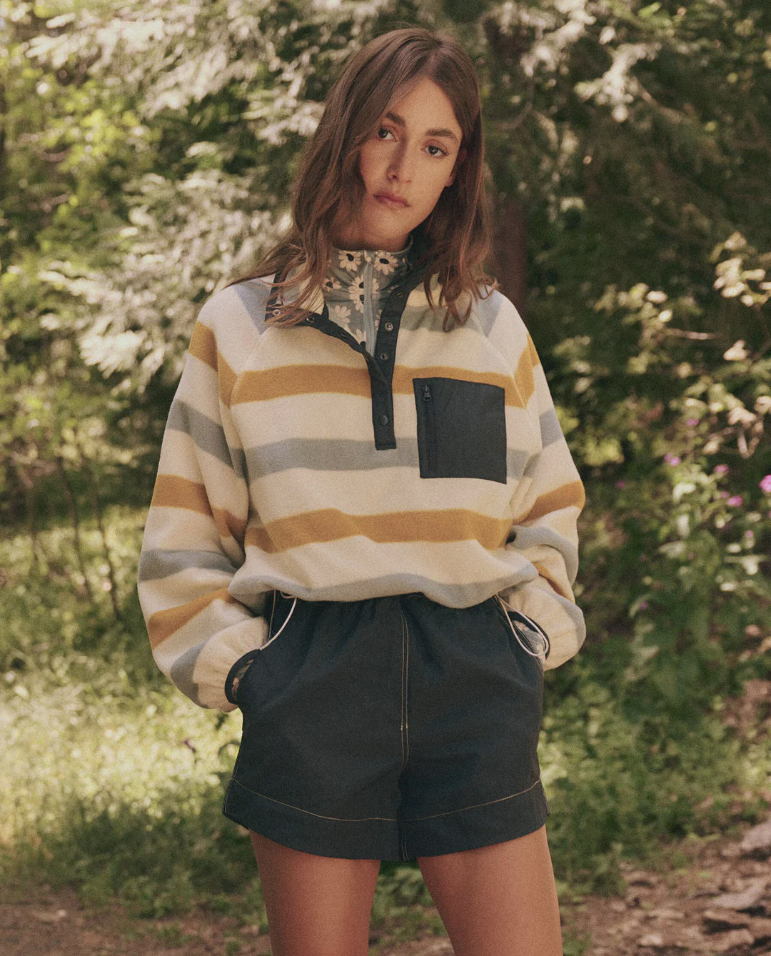 A young woman stands outdoors, wearing The Excursion Pullover from The Great Inc., along with dark shorts. Her hands are in her pockets, and she has long, wavy hair. Lush green trees create a forest backdrop.