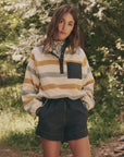 A young woman stands outdoors, wearing The Excursion Pullover from The Great Inc., along with dark shorts. Her hands are in her pockets, and she has long, wavy hair. Lush green trees create a forest backdrop.