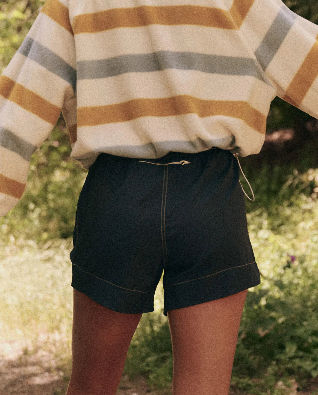 A person stands outdoors with their back to the camera, wearing a long-sleeved top with yellow, gray, and white stripes paired with The Rover Short by The Great Inc., featuring dark water-repellent fabric. The blurred greenery in the background enhances the natural setting.