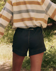 A person stands outdoors with their back to the camera, wearing a long-sleeved top with yellow, gray, and white stripes paired with The Rover Short by The Great Inc., featuring dark water-repellent fabric. The blurred greenery in the background enhances the natural setting.