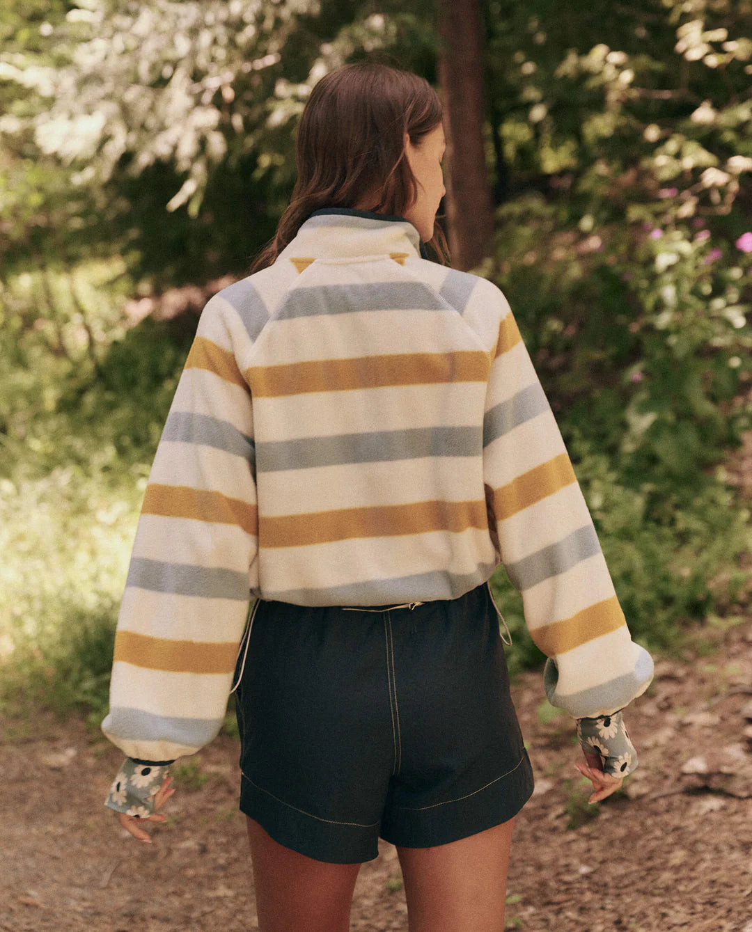 Wearing The Great Inc.'s Excursion Pullover, a person with long hair and dark shorts navigates a dirt path through a wooded area. The lightweight striped fleece jacket in cream, yellow, and blue complements the sunny skies and vivid green foliage of their daily adventure.