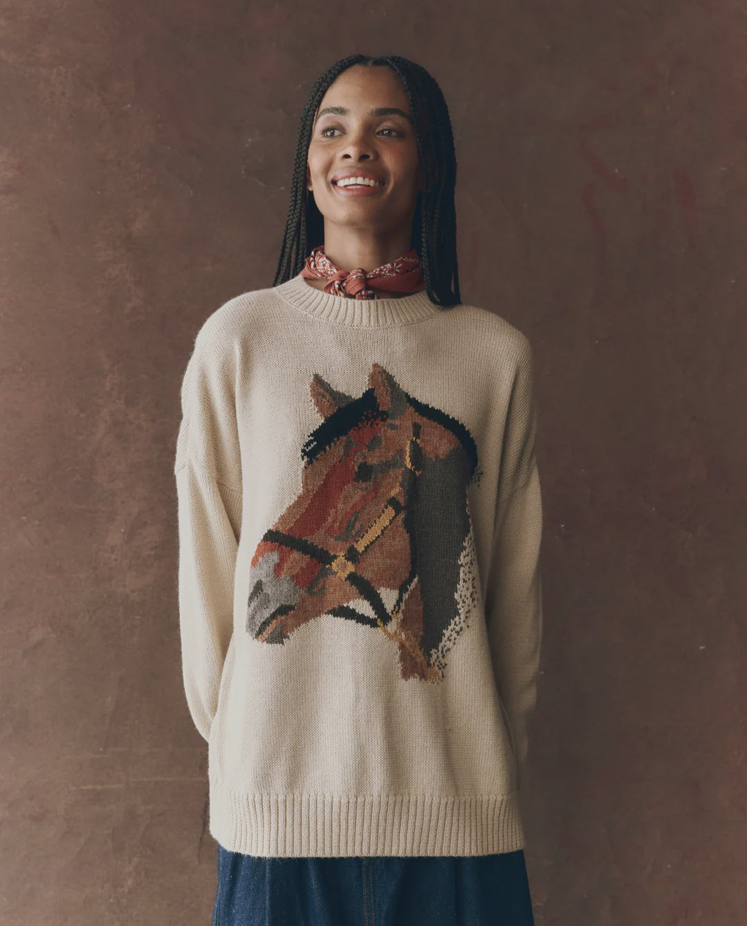 A woman with braided hair smiles in The Great Inc. Stable Pullover, an oversized crew sweater with a large horse intarsia graphic. She pairs it with a red patterned neck scarf, standing against a brown background.