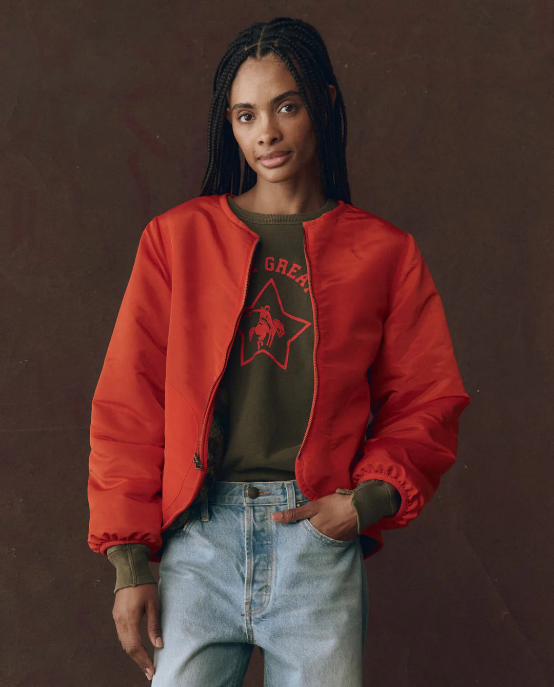 A woman with long braided hair wears The Great Inc. Reversible Plush Bomber in bright red over a green graphic sweatshirt and light blue jeans, standing against a plain dark background.