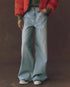A person wearing The Great Inc. Slouch Wide Leg jean in light blue, a green top, red jacket, and white sneakers stands against a plain brown background with hands in pockets and face not visible.
