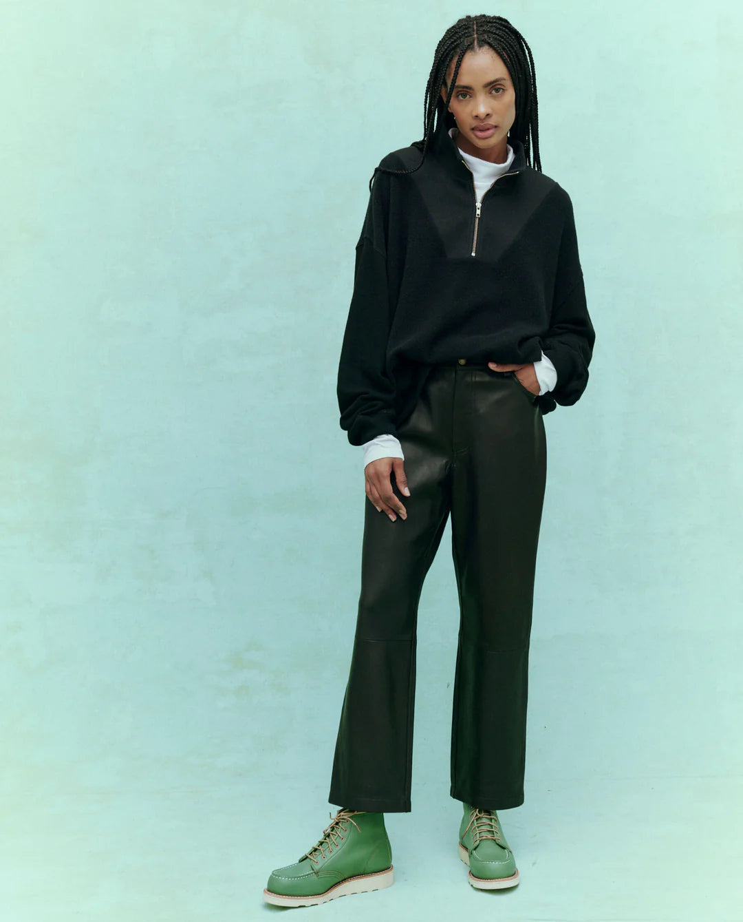 Against a light teal background, a person with braided hair stands confidently, showcasing style in The Great Inc.&#39;s The Leather Wayne. They wear a black sweater, white turtleneck, and soft leather pants paired with green boots, one hand casually in their pocket—capturing the essence of Wayne Jean flair.