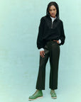 Against a light teal background, a person with braided hair stands confidently, showcasing style in The Great Inc.'s The Leather Wayne. They wear a black sweater, white turtleneck, and soft leather pants paired with green boots, one hand casually in their pocket—capturing the essence of Wayne Jean flair.