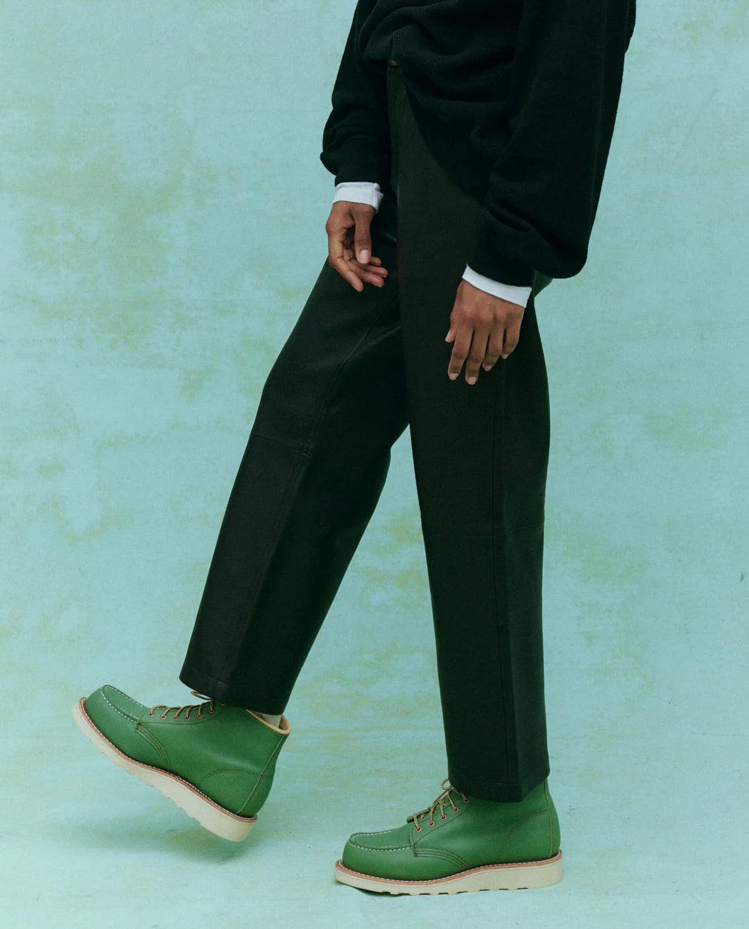 A person is posing against a light green background, wearing a black outfit and The Great Inc.'s soft leather "The Leather Wayne" green lace-up boots. Their left leg is slightly raised, emphasizing the slouchy body of the footwear.