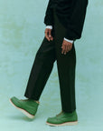 A person is posing against a light green background, wearing a black outfit and The Great Inc.'s soft leather "The Leather Wayne" green lace-up boots. Their left leg is slightly raised, emphasizing the slouchy body of the footwear.