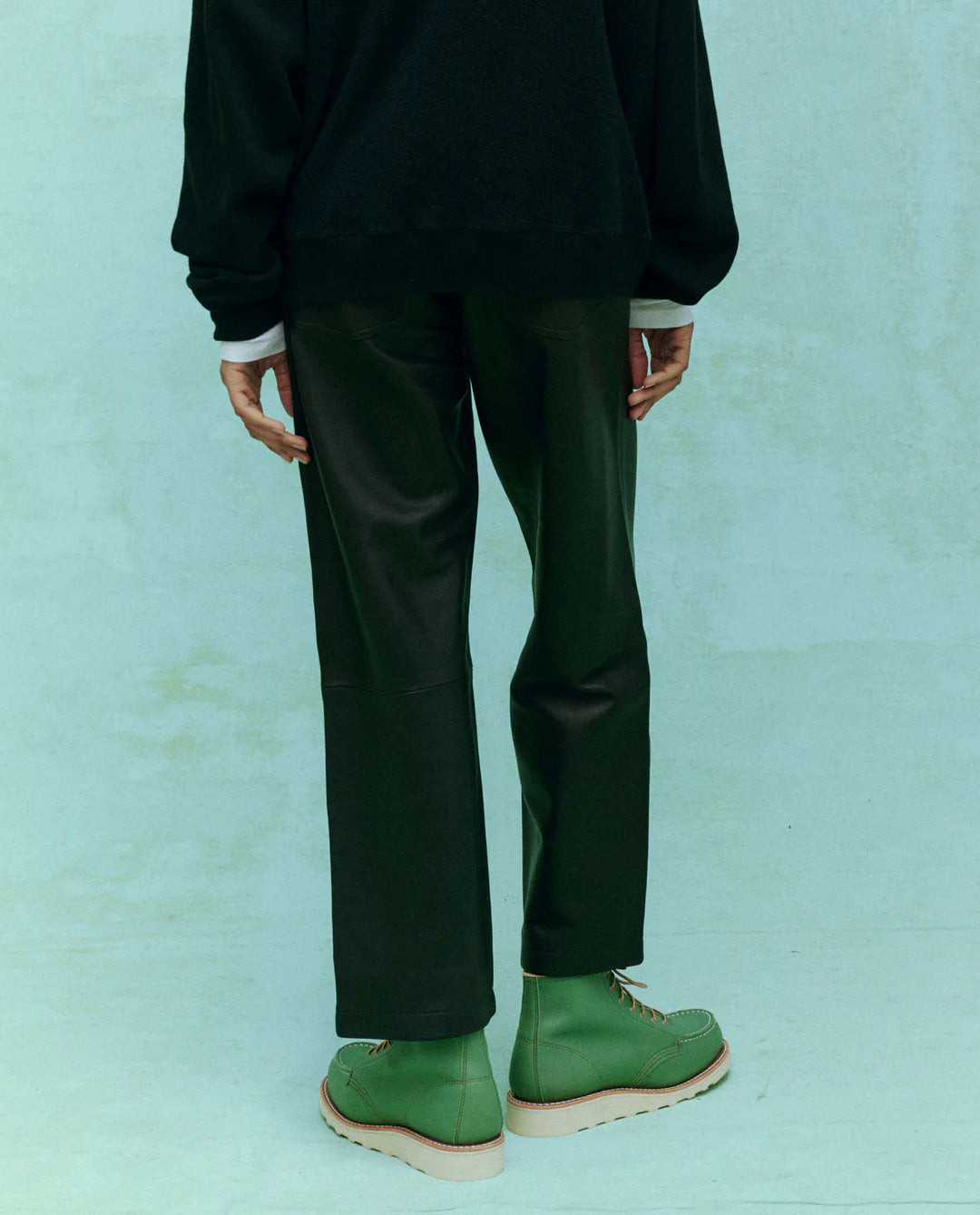 Wearing &quot;The Leather Wayne&quot; by The Great Inc., a person stands against a light green background in black sweatshirt and pants, highlighting their relaxed silhouette. Green boots with beige soles contrast the ensemble, while the photo captures the full outfit from behind, seamlessly blending soft leather elements with effortless style.