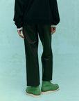 Wearing "The Leather Wayne" by The Great Inc., a person stands against a light green background in black sweatshirt and pants, highlighting their relaxed silhouette. Green boots with beige soles contrast the ensemble, while the photo captures the full outfit from behind, seamlessly blending soft leather elements with effortless style.