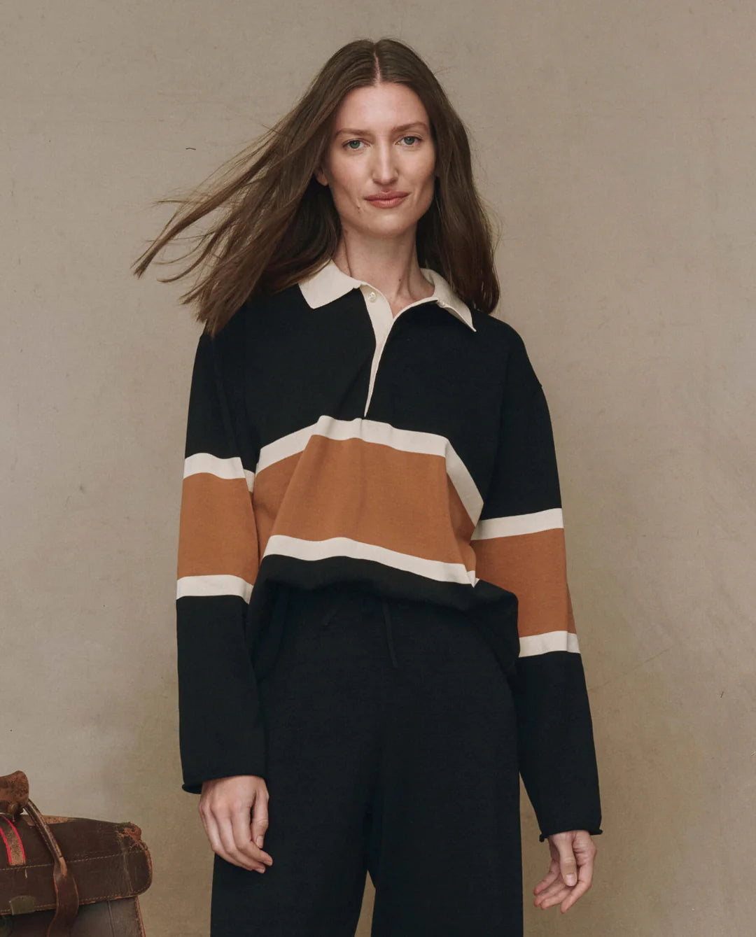 A woman with long brown hair wears The Rugby Sweater by The Great Inc.—a boxy knit black pullover with wide brown and white stripes—standing against a plain beige background, looking at the camera and smiling slightly.