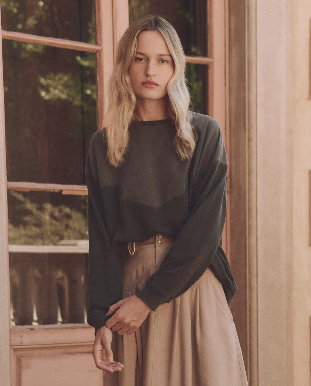 A woman with long blonde hair wears The Great Inc.'s Western Slouch Sweatshirt and a beige skirt, standing by a large window. She gazes at the camera, exuding elegance in the soft light and wooden door—a perfect vintage find.