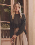 A woman with long blonde hair wears The Great Inc.'s Western Slouch Sweatshirt and a beige skirt, standing by a large window. She gazes at the camera, exuding elegance in the soft light and wooden door—a perfect vintage find.