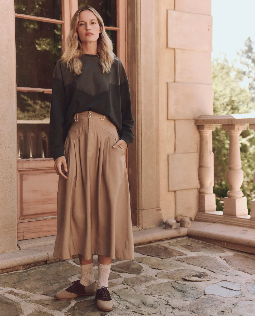 A person with long hair stands on a stone patio by a building with large windows, wearing The Western Slouch Sweatshirt by The Great Inc. and beige wide-leg pants, paired with socks and brown shoes. Trees in the background enhance the handcrafted Los Angeles vibe.