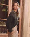 A long-haired blonde woman stands in front of a glass door, wearing The Great Inc.'s dark Western Slouch Sweatshirt and light pants, posing casually with one hand in her pocket against a softly lit background with muted, earthy Los Angeles-inspired tones.