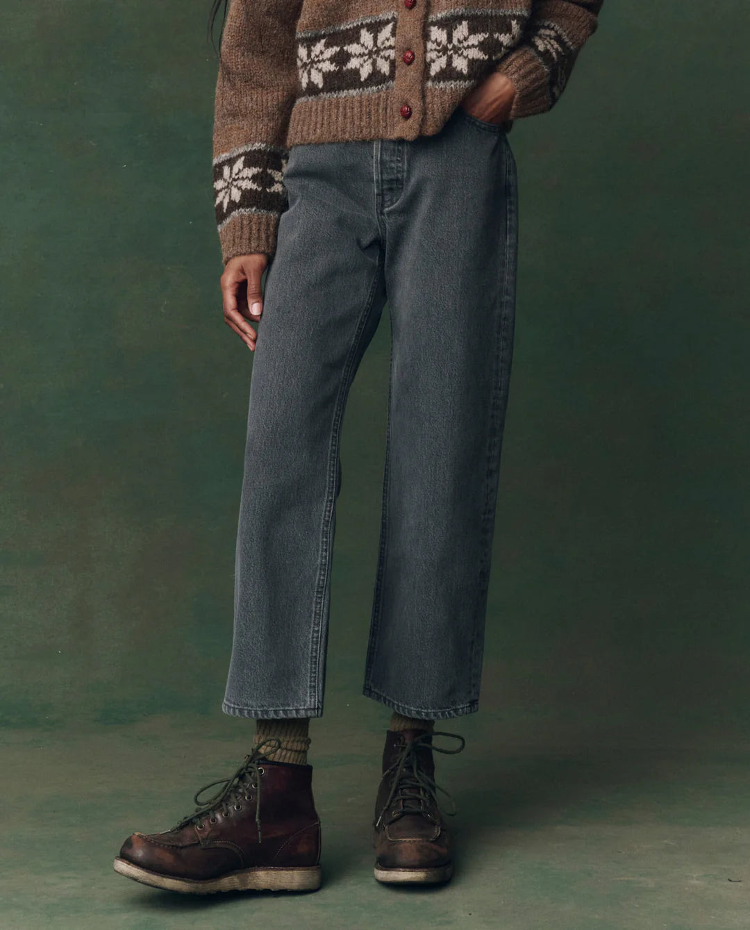 Against a green background, a person stands with hands in their pockets wearing The Wayne Jean by The Great Inc., paired with a brown patterned sweater and dark brown lace-up boots. Their face is not visible.