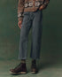 Against a green background, a person stands with hands in their pockets wearing The Wayne Jean by The Great Inc., paired with a brown patterned sweater and dark brown lace-up boots. Their face is not visible.