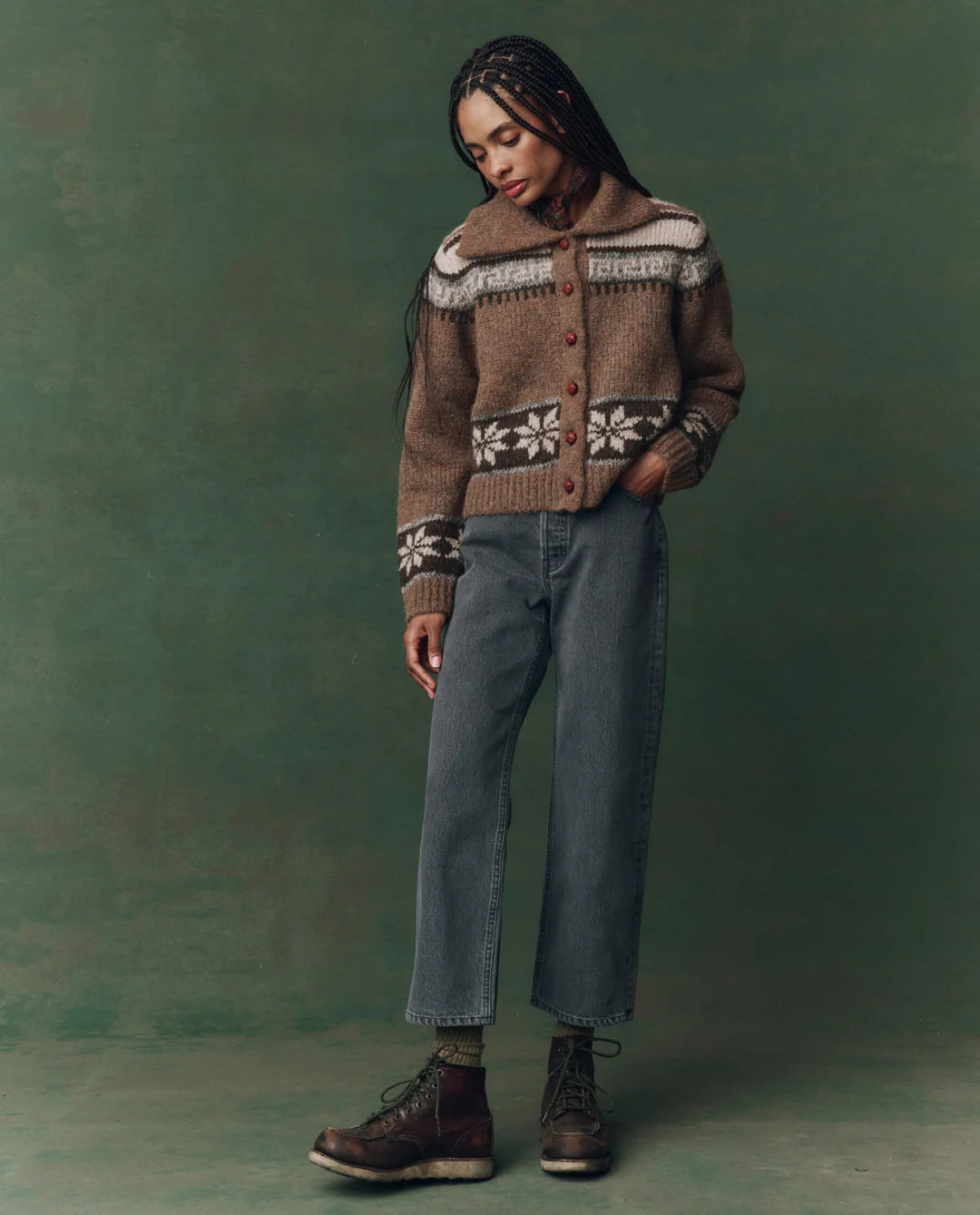 A person with long braids stands against a green background, wearing The Wayne Jean by The Great Inc., paired with a brown and white patterned sweater and brown lace-up boots. One hand in their pocket, they embody LA's effortless style.