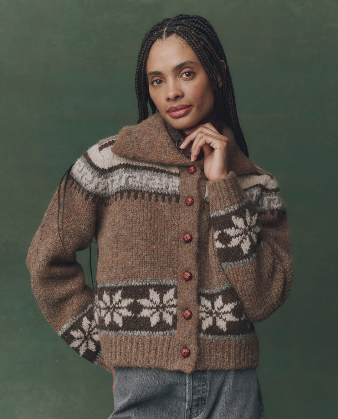 A person with long braids poses with one hand near their chin against a green background, wearing The Great Inc.’s The Nordic Scholar Cardigan—an oversized knit sweater featuring white floral designs and red buttons.