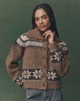 A person with long braids poses with one hand near their chin against a green background, wearing The Great Inc.’s The Nordic Scholar Cardigan—an oversized knit sweater featuring white floral designs and red buttons.