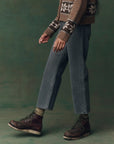 A person wears The Wayne Jean by The Great Inc., paired with a brown patterned sweater, olive socks, and dark brown lace-up boots. Only their lower half is visible against a green background.