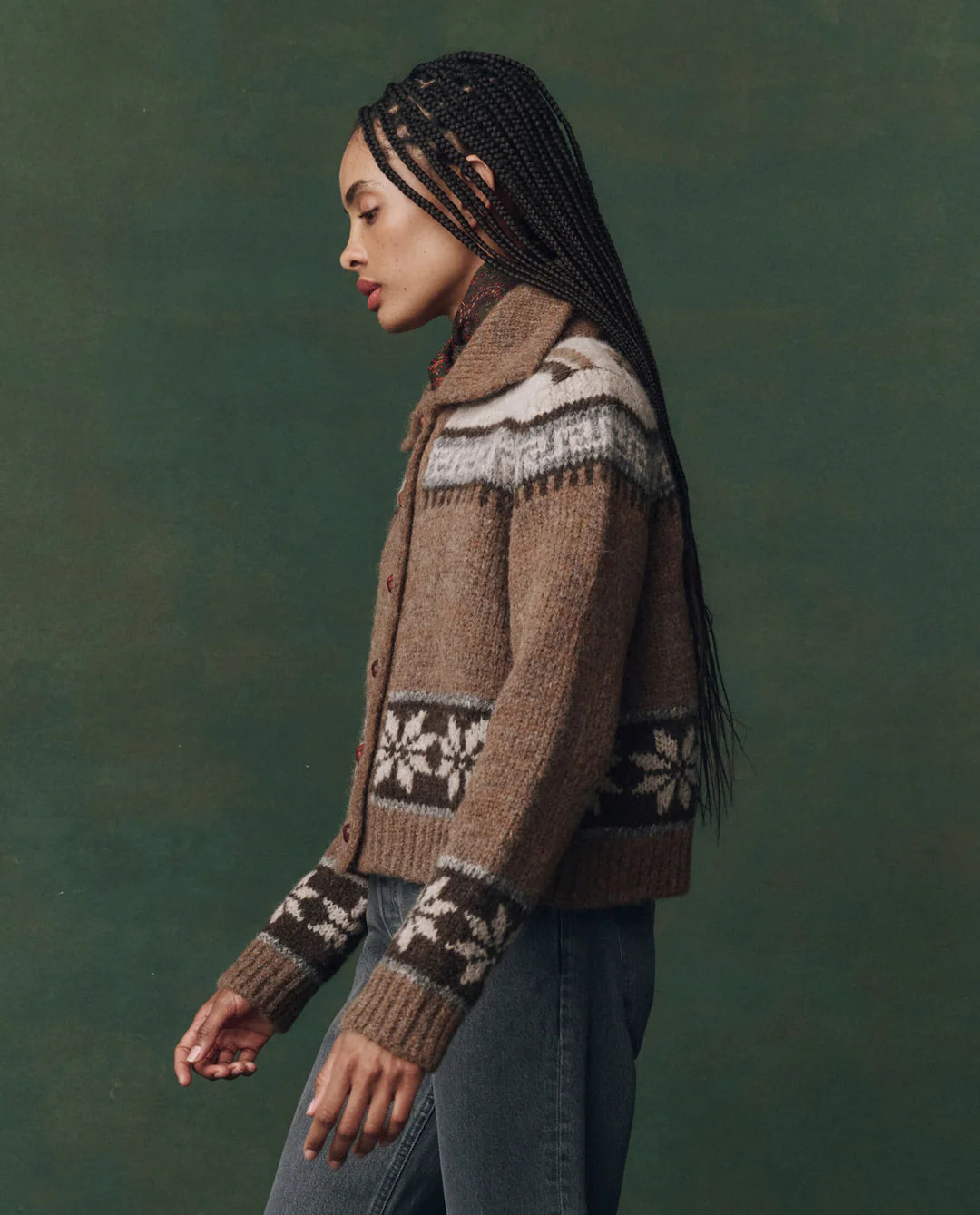 A woman with long braided hair wears The Nordic Scholar Cardigan by The Great Inc. in brown, paired with blue jeans, standing in profile against a green background.