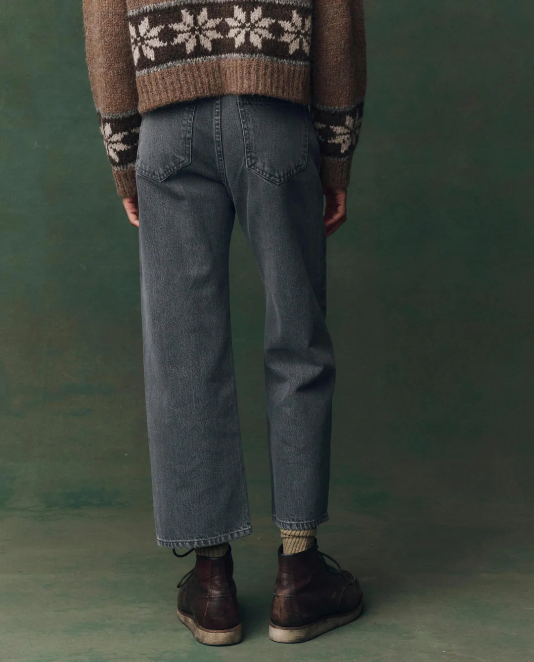 A person faces away, wearing The Wayne Jean by The Great Inc., paired with a brown sweater featuring white snowflakes and dark brown lace-up boots, standing against a green studio backdrop.