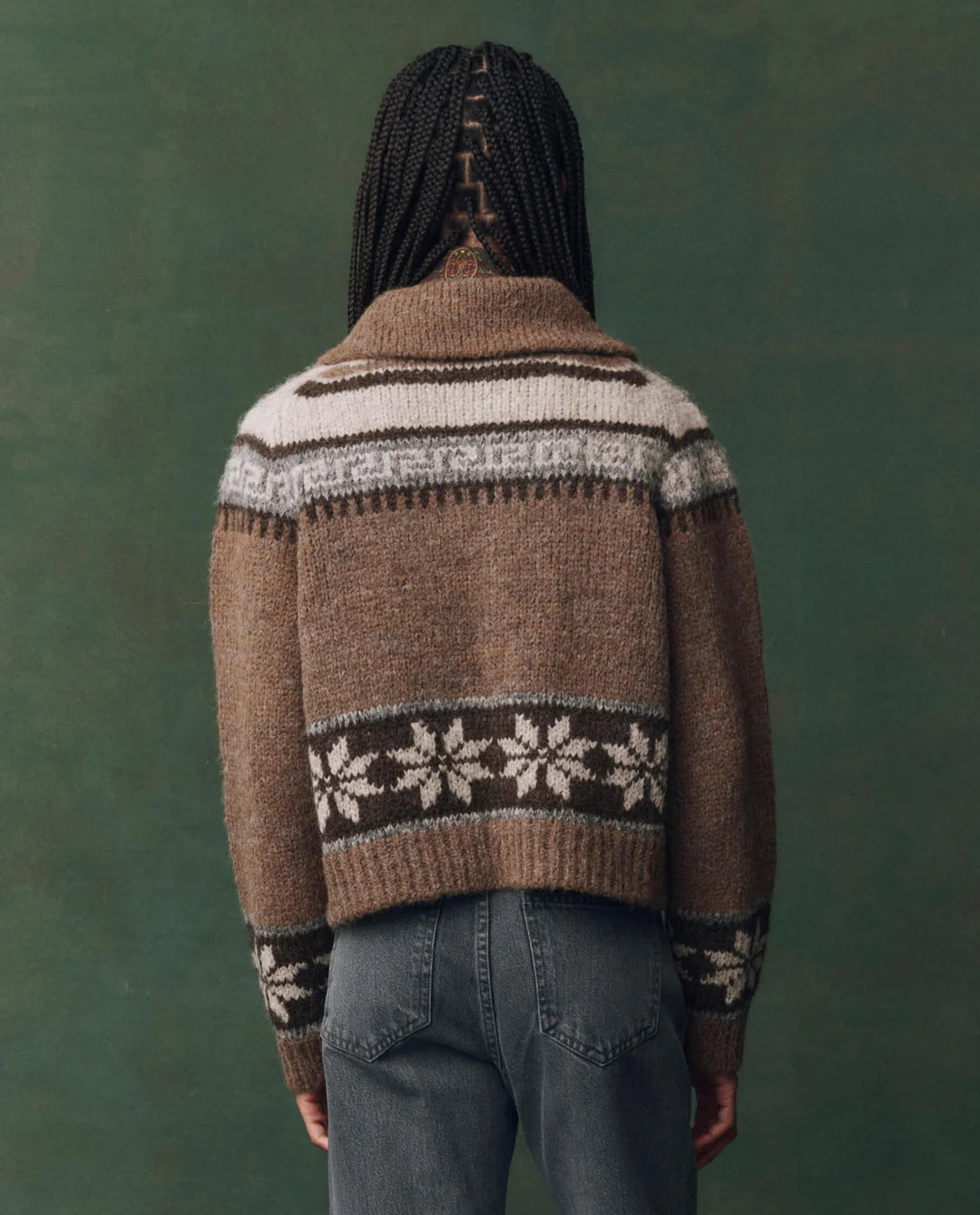 A person with braided hair, seen from behind, wears The Great Inc.'s Nordic Scholar Cardigan—brown with white and dark geometric snowflake patterns—paired with blue jeans, set against a muted green background.