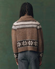 A person with braided hair, seen from behind, wears The Great Inc.'s Nordic Scholar Cardigan—brown with white and dark geometric snowflake patterns—paired with blue jeans, set against a muted green background.