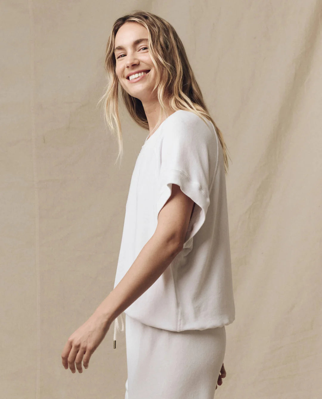 A woman with long blonde hair smiles at the camera, wearing The Wedge Sweatshirt by The Great Inc. and matching white bottoms, posed against a neutral beige background.